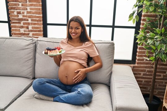 Young latin woman pregnant eating sweets sitting on sofa at home