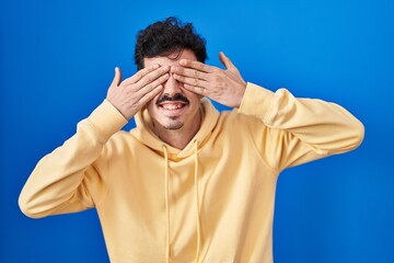 Hispanic man standing over blue background covering eyes with hands smiling cheerful and funny. blind concept.