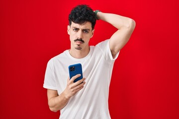 Hispanic man using smartphone over red background confuse and wondering about question. uncertain with doubt, thinking with hand on head. pensive concept.