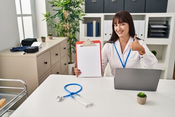 Young brunette doctor woman holding clipboard smiling happy and positive, thumb up doing excellent and approval sign