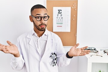 African american optician man standing by eyesight test clueless and confused expression with arms and hands raised. doubt concept.