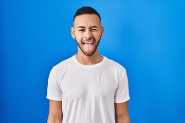 Young hispanic man standing over blue background sticking tongue out happy with funny expression. emotion concept.
