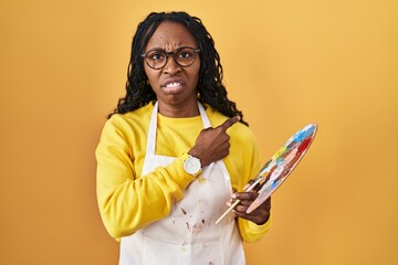 African woman holding painter palette pointing aside worried and nervous with forefinger, concerned and surprised expression