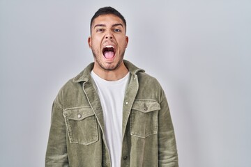 Fototapeta premium Young hispanic man standing over isolated background angry and mad screaming frustrated and furious, shouting with anger. rage and aggressive concept.