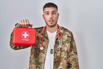 Young hispanic doctor wearing camouflage army uniform holding first aid kit scared and amazed with...