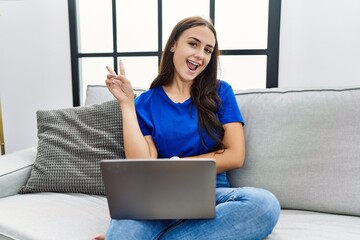 Fototapeta na wymiar Young brunette woman using laptop at home smiling with happy face winking at the camera doing victory sign. number two.