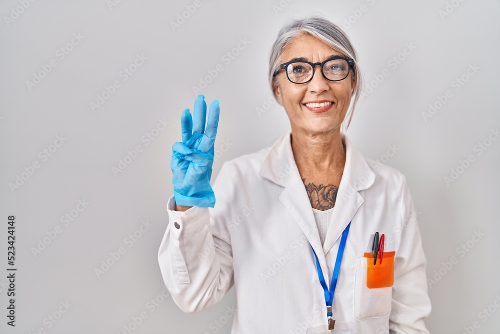 Poster middle age woman with grey hair wearing scientist robe showing and pointing up with fingers number t