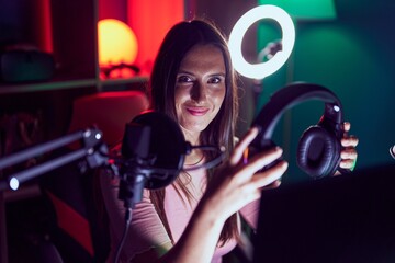 Young beautiful hispanic woman streamer smiling confident holding headphones at gaming room