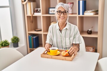 Middle age grey-haired woman having breakfast sitting on table at home