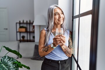 Middle age grey-haired woman smiling confident drinking water at home