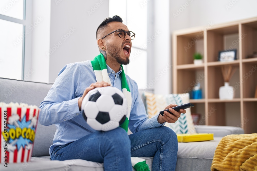 Sticker Young hispanic man football hooligan holding ball supporting team angry and mad screaming frustrated and furious, shouting with anger looking up.