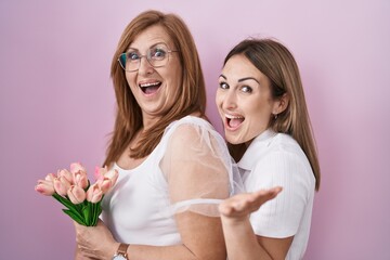 Hispanic mother and daughter holding pink tulips bouquet celebrating achievement with happy smile and winner expression with raised hand