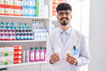 Young hispanic man pharmacist smiling confident holding pills bottles at pharmacy