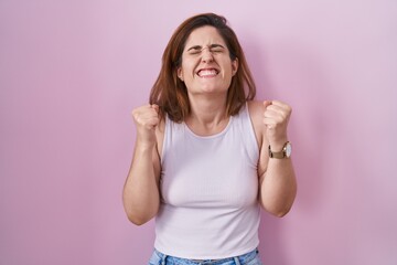 Obraz na płótnie Canvas Brunette woman standing over pink background excited for success with arms raised and eyes closed celebrating victory smiling. winner concept.