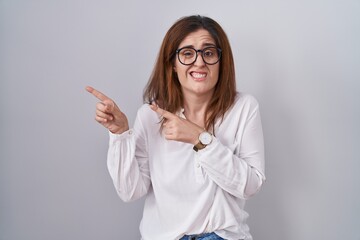 Brunette woman standing over white isolated background pointing aside worried and nervous with both hands, concerned and surprised expression