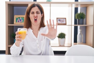 Brunette woman drinking glass of orange juice doing stop gesture with hands palms, angry and frustration expression