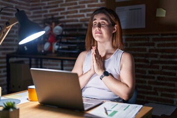 Brunette woman working at the office at night praying with hands together asking for forgiveness...