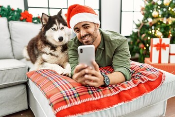 Young hispanic man make selfie by the smartphone lying on sofa with dog by christmas tree at home