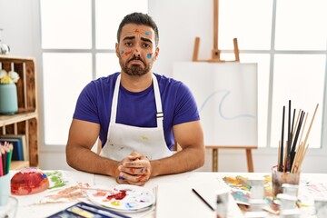 Young hispanic man with beard at art studio with painted face depressed and worry for distress,...