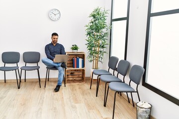 Young hispanic man using laptop sitting on chair at waiting room