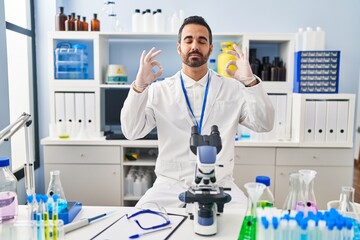 Young hispanic man with beard working at scientist laboratory relax and smiling with eyes closed doing meditation gesture with fingers. yoga concept.