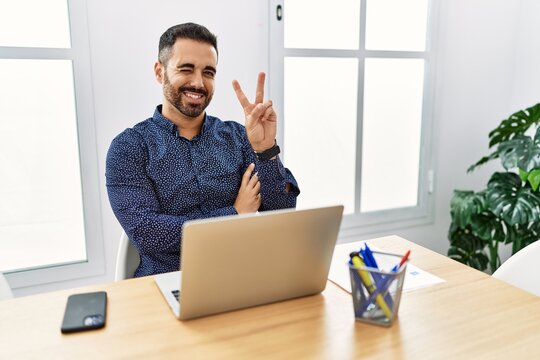 Young hispanic man with beard working at the office with laptop smiling with happy face winking at the camera doing victory sign with fingers. number two.