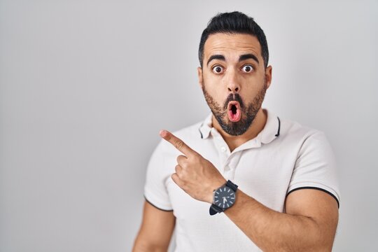 Young hispanic man with beard wearing casual clothes over white background surprised pointing with finger to the side, open mouth amazed expression.
