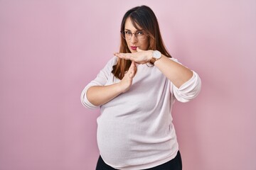 Pregnant woman standing over pink background doing time out gesture with hands, frustrated and...
