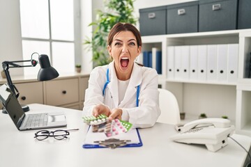 Young brunette doctor woman holding pills angry and mad screaming frustrated and furious, shouting with anger. rage and aggressive concept.