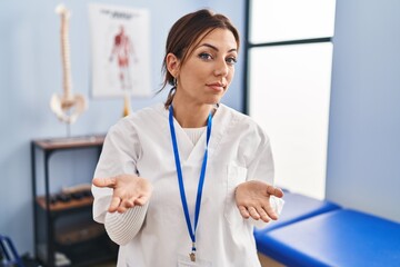 Young brunette woman working at pain recovery clinic clueless and confused with open arms, no idea...