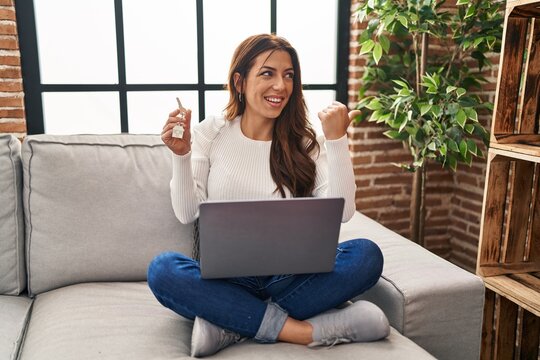 Young brunette woman using laptop holding keys of new home pointing thumb up to the side smiling happy with open mouth