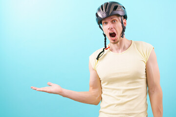 A shocked man with an open mouth wearing a bicycle helmet points with his hand at an empty copy space on a blue background. Lifestyle. Looking. Male. Object. Person. Protection. Road. Studio