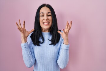 Hispanic woman standing over pink background celebrating mad and crazy for success with arms raised...