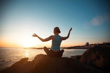 A middle-aged woman does yoga, meditating on the ocean beach during a beautiful sunset.