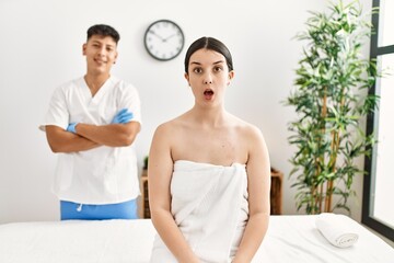 Obraz na płótnie Canvas Young woman at wellness spa with professional therapist sitting on massage table scared and amazed with open mouth for surprise, disbelief face