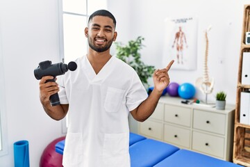 Naklejka na ściany i meble Young indian physiotherapist holding therapy massage gun at wellness center with a big smile on face, pointing with hand finger to the side looking at the camera.