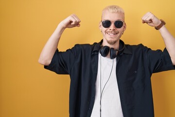 Young caucasian man wearing sunglasses standing over yellow background showing arms muscles smiling proud. fitness concept.
