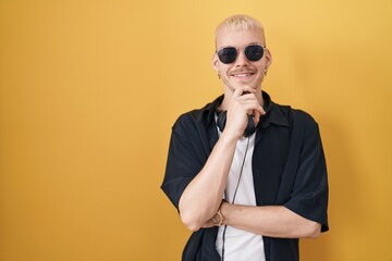 Young caucasian man wearing sunglasses standing over yellow background with hand on chin thinking about question, pensive expression. smiling and thoughtful face. doubt concept.