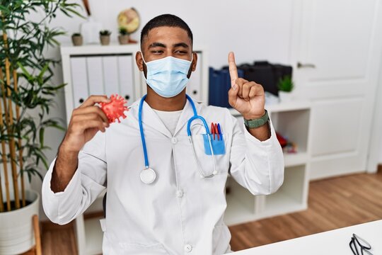 Young indian doctor holding virus toy wearing safety mask smiling with an idea or question pointing finger with happy face, number one