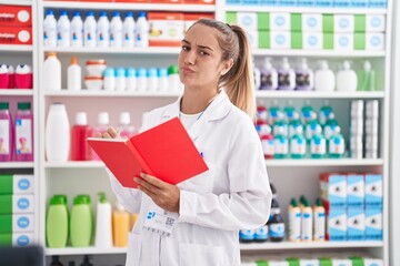 Young blonde woman working at pharmacy drugstore holding notebook smiling looking to the side and staring away thinking.