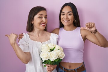 Fototapeta premium Hispanic mother and daughter holding bouquet of white flowers pointing to the back behind with hand and thumbs up, smiling confident