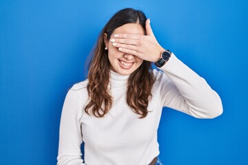 Young hispanic woman standing over blue background smiling and laughing with hand on face covering...