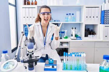 Young hispanic woman working at scientist laboratory shouting with crazy expression doing rock...