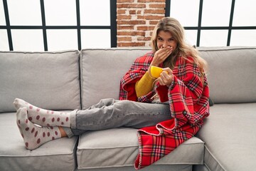 Young woman sitting on the sofa drinking a coffee at home laughing and embarrassed giggle covering...