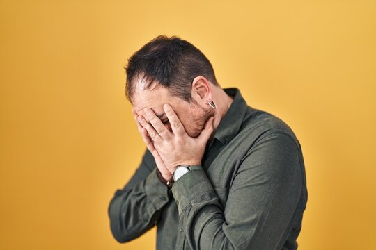 Plus size hispanic man with beard standing over yellow background with sad expression covering face with hands while crying. depression concept.