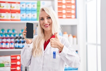 Caucasian woman working at pharmacy drugstore showing smartphone screen smiling happy and positive, thumb up doing excellent and approval sign