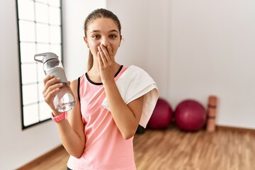 Young brunette teenager wearing sportswear holding water bottle laughing and embarrassed giggle...