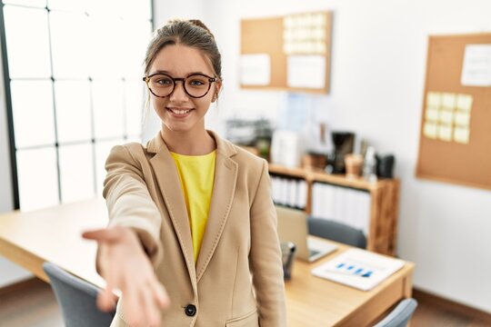 Young brunette teenager wearing business style at office smiling friendly offering handshake as greeting and welcoming. successful business.