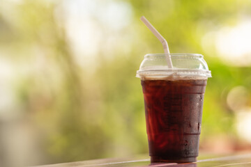 Closeup of takeaway plastic cup of iced black coffee Americano on wooden table with green nature background.
