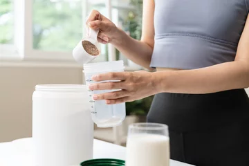 Poster Young sporty woman pouring protein powder into a cup to make replacement food meal after workout © Pormezz